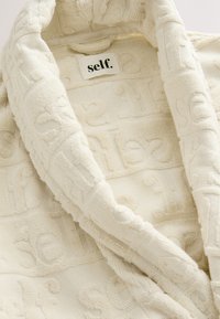Robe blanche texturée présentant un motif en relief du mot « self. » sur le tissu, avec une étiquette noire au niveau de l'encolure.