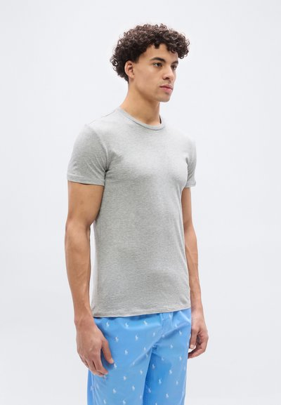 Νεαρός άνδρας με σγουρά μαλλιά που φοράει εφαρμοστό γκρι μπλουζάκι και ανοιχτόχρωμο μπλε σορτς με λευκά σχέδια αλόγου, στέκεται μπροστά σε απλό φόντο.