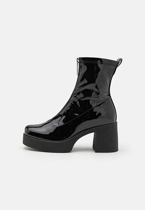 Botas de tobillo de charol negro con cremallera lateral, punta redondeada y un tacón grueso y texturizado. El diseño cuenta con un acabado suave y brillante.