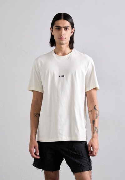 T-shirt crème clair en tissu doux avec des manches courtes, arborant un logo noir "MSGM" centré sur la poitrine; associé à un short noir déchiré.