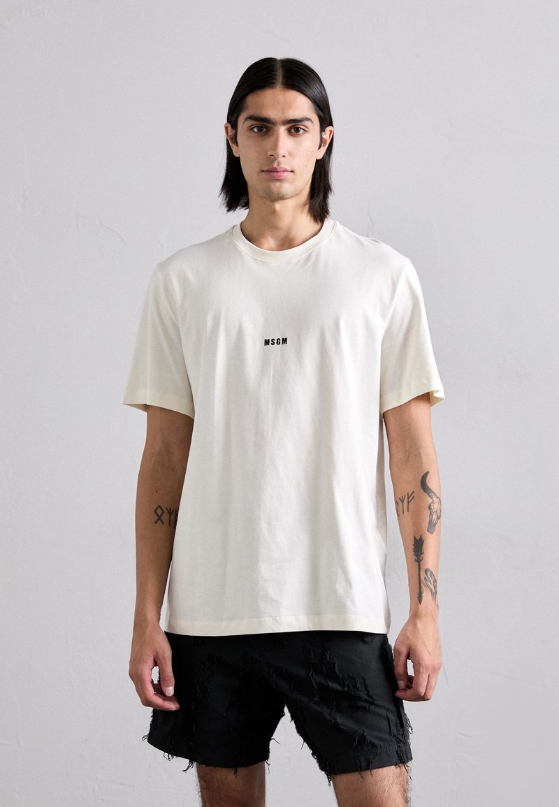 Lys kremfarget t-skjorte i myk stoff med korte ermer, med en svart "MSGM" logo sentrert på brystet; parret med slitte svarte shorts.
