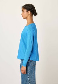 Mėlynas ilgomis rankovėmis megztinis su tekstūruotu paviršiumi ir apvaliu iškirpimu, derinamas su šviesiai mėlynomis džinsais.
