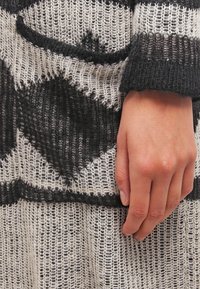 Vêtement en maille à motifs noirs et crème avec un tissage lâche. Présente des motifs géométriques et des poignets texturés. Main posée sur le tissu.