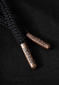 Cordones de zapatos negros trenzados con puntas metálicas de bronce grabadas con el texto "Reell", sobre un fondo de tela oscura.