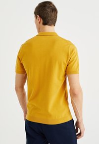 Short-sleeve polo shirt in mustard yellow, textured knit fabric, with a traditional collar and ribbed cuffs, fitted design.