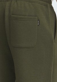 Olivenfargede joggebukser laget av myk stoff med en glatt tekstur. Funksjoner inkluderer en lappelomme og en liten merkeetikett på siden.