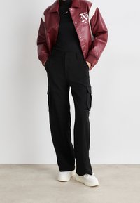 Burgunderfarbene Leder-Collegejacke mit weißen Akzenten, schwarze Cargo-Hosen mit Seitentaschen und weiße Sneakers, die vor einem neutralen Hintergrund stehen.