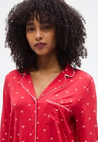 Κόκκινο πουκάμισο από απαλό ύφασμα με μοτίβα λευκού φιόγκου, γιακά με κόψιμο και τσέπη με λεπτομέρειες λευκής τρέσας.