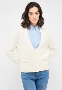 Kofta i off-white med v-ringning, fem knappar och två framfickor. Bärs över en ljusblå randig skjorta och jeans.