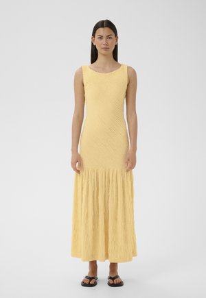 Kvinde iført en ærmeløs, tekstureret gul maxikjole og sorte sandaler, stående mod en ensfarvet lys grå baggrund.