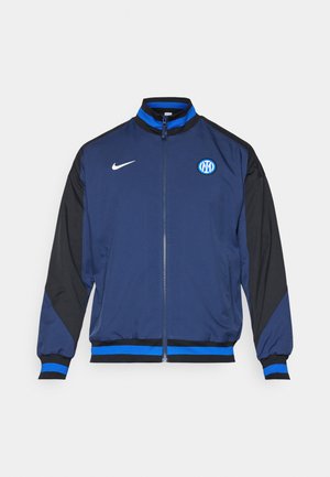 Chaqueta azul marina con mangas negras, que cuenta con un cierre completo, puños acanalados, detalles en azul y un logo circular en la parte izquierda del pecho.