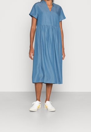 Kvinde iført en løs blå midikjole med korte ærmer og hvide sneakers med gule detaljer, stående op ad en ensfarvet væg.