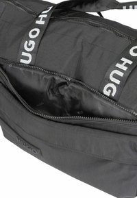 Borsa di tessuto nera di forma rettangolare, con apertura a zip, dettaglio del logo in gomma e doppi manici con il marchio "HUGO".