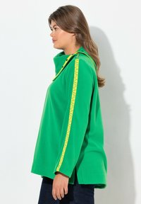 Suéter verde con un cuello alto, que presenta una franja amarilla con un patrón a lo largo de las mangas y detalles de botones en los bordes del cuello.