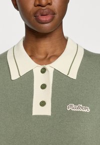 Zaļš rievots polo krekls ar krēmkrāsas apkakli ar zaļām svītrām, trim zaļiem pogām un izšūtu zīmola logo uz krūtīm.