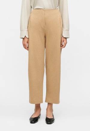 Γυναίκα που φοράει μπεζ παντελόνι με ίσια μπατζάκια, κρεμ πουκάμισο με μακριά μανίκια και μαύρα παπούτσια με τετράγωνη μύτη, στέκεται μπροστά σε λευκό φόντο.