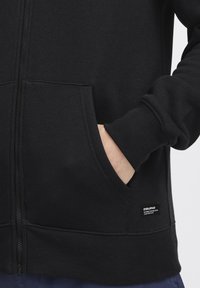 Zwarte rits hoodie gemaakt van zachte stof, met geribde manchetten en zoom, zijzakken en een klein merklabel op de onderkant rechts.