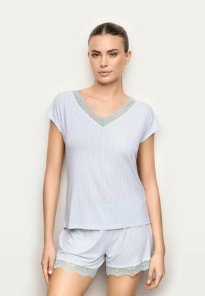 Kobieta ubrana w jasnoniebieską top na krótki rękaw w paski i dopasowane szorty z koronkowym wykończeniem, stojąca na tle jednolitego jasnego tła.