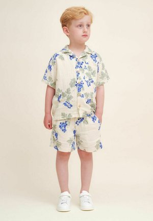 Kortærmet skjorte med knap og shorts sæt i creme med grønne og blå drue mønstre, lavet af letvægtsstof. Hvide sneakers.