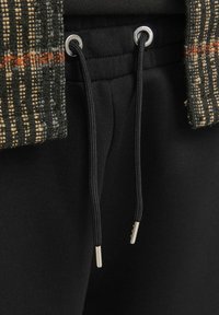 Pantaloni jogger negri cu o textură netedă, prevăzuți cu șnururi cu capete metalice și un brâu din țesătură cu model în tonuri închise.