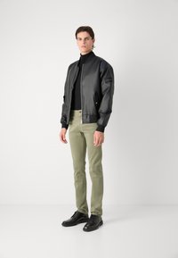 Černá kožená bunda ve stylu bomber s ribbeny na rukávech, zipovým zapínáním a bočními kapsami, v kombinaci s olivově zelenými kalhotami a černými šněrovacími botami.