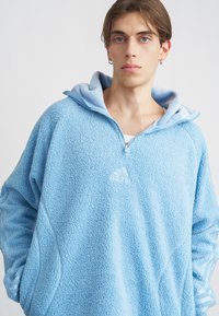 Giovane con i capelli castano chiaro che indossa un pullover in pile azzurro chiaro con una mezza zip e cappuccio, in piedi davanti a uno sfondo semplice.