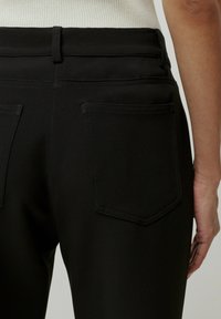 Zwarte broek van gladde stof. Beschikt over een tailleband, twee achterzakken en subtiele stiksels. Textuur lijkt soepel en aansluitend.