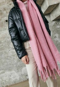 Personne portant une doudoune noire, une longue écharpe rose pâle douce et un pantalon crème, debout contre un mur de béton texturé.