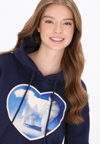 Sudadera con capucha azul marino con un gráfico en forma de corazón de un gato blanco sobre un fondo azul. Cuenta con capucha con cordón y textura de tela suave.