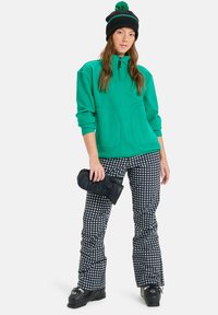 Pullover de fleece verde com gola de fecho, dois bolsos frontais; calças impermeáveis preto e branco com bolinhas; gorro preto de malha com pom-pom, segurando luvas.