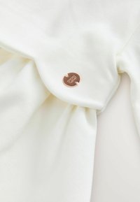 Weißer Stoff mit einer weichen Textur, der mit einem kleinen, kupferfarbigen Knopf, der ein "B" eingeprägt hat, verziert ist und an einem dünnen weißen Faden befestigt ist.