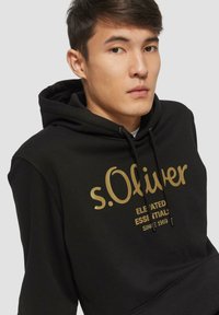 Zwarte hoodie met een gouden bedrukte logo "s.Oliver" en tekst. Gemaakt van zachte, gestructureerde stof met een kangoeroe zak en een capuchon met trekkoord.
