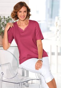 Korte mouw, V-hals shirt in fuchsia met decoratieve studs op de halslijn. Gecombineerd met witte, cropped broeken, tegen een heldere achtergrond.