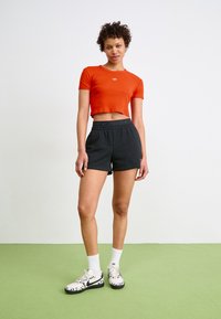 Kuttet rød t-skjorte med logo, kombinert med svarte shortser med elastisk midjebånd. Fottøy: hvite joggesko med svarte detaljer og mønster.