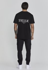 Čierne bavlnené tričko s bielym logom "SIKSILK" na chrbte, v kombinácii s čiernymi cargo nohavicami s bočnými vreckami a bielymi teniskami.