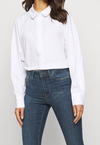 Hvid skjorte med krave, knebelukning og lange ærmer, kombineret med blå denimjeans med forlomme og standard pasform.