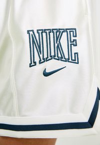 Tejido blanco con texto bordado en azul marino "NIKE" y logotipo de swoosh por encima del dobladillo, con detalle de ribete en azul marino.