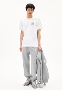 Witte katoenen t-shirt met een bedrukte print, gecombineerd met grijzige fleece joggingsbroek met een wit embleem. Model houdt een grijze sweatshirt vast.