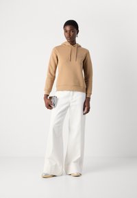 Felpa beige con tasca a marsupio e coulisse, abbinata a pantaloni bianchi a gamba larga. Borsa argentata che completa l'outfit.