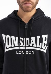 Sudadera con cremallera negra con el logo en blanco "LONSDALE LONDON" en el frente. Cuenta con cordones, una textura suave y un ajuste regular.