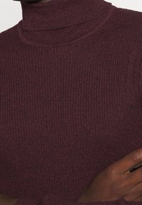 Bordeauxfarbener gerippter Rollkragenpullover aus strukturiertem Stoff. Nahaufnahme zeigt den hohen Kragen und das taillierte Design mit sichtbaren Nähdetails.