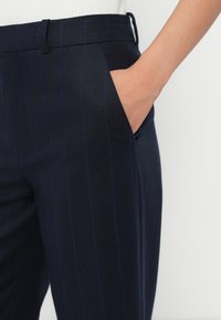 Navy trousers with light pinstripes, featuring a side pocket and seamless waistband, made from a smooth fabric with a tailored fit.