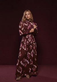 Robe longue fleurie couleur bordeaux profond avec des fleurs roses et blanches. Elle présente des manches bouffantes, une taille cintrée et des volants superposés à l'ourlet.