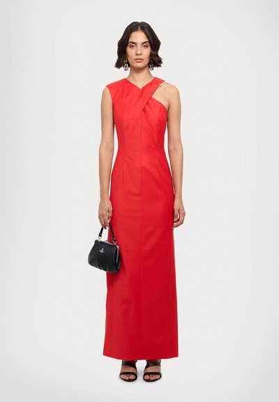Robe longue rouge, sans manches, près du corps, avec un décolleté asymétrique et des détails plissés. Associée à un petit sac à main noir et des sandales noires.