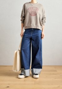Sudadera gris con gráfico rojo, pantalones anchos de mezclilla azul con bajos deshilachados y zapatillas negras y blancas. La modelo sostiene un bolso tote beige.