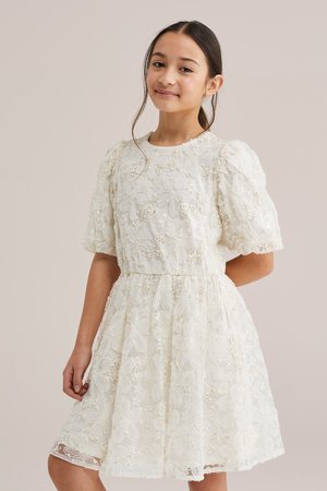 Jeune fille aux cheveux foncés attachés en une queue de cheval basse, portant une robe blanche en dentelle florale avec des manches courtes bouffantes, posant devant un fond uni.