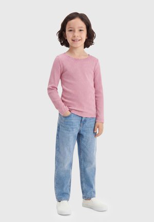 Smilende barn med kort mørkt hår står med den ene hånd i lommen iført pink langærmet trøje, lyseblå jeans og hvide slip-on sko.