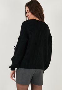 Fekete bordázott pulóver, amely kényelmes oversized szabású és ejtett vállú. A ujjakon kis fekete masnik találhatók. Szürke sorttal párosítva.