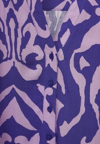 Koszula z guzikami w głębokim fioletowym i jasnym lawendowym kolorze z odważnym, abstrakcyjnym wzorem. Posiada dekolt w kształcie litery V oraz pięć pasujących guzików z przodu.