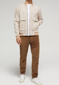 Light beige bomber jacket with a textured finish, two front pockets, ribbed cuffs, and a contrasting collar. Brown corduroy pants and white sneakers.
